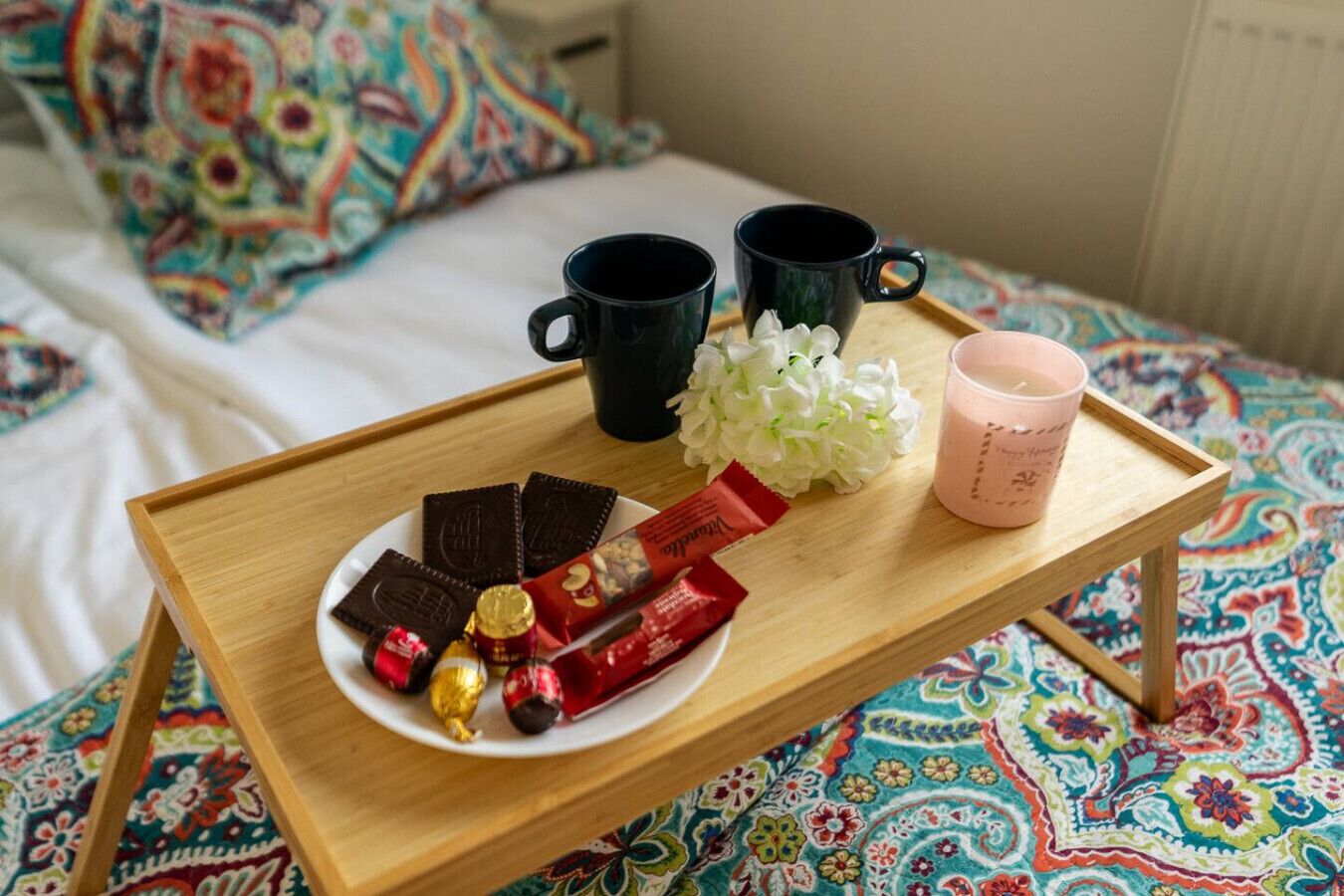 Una bandeja con bombones, una vela rosa y dos tazas negras sobre una cama con una colcha de diseño colorido.