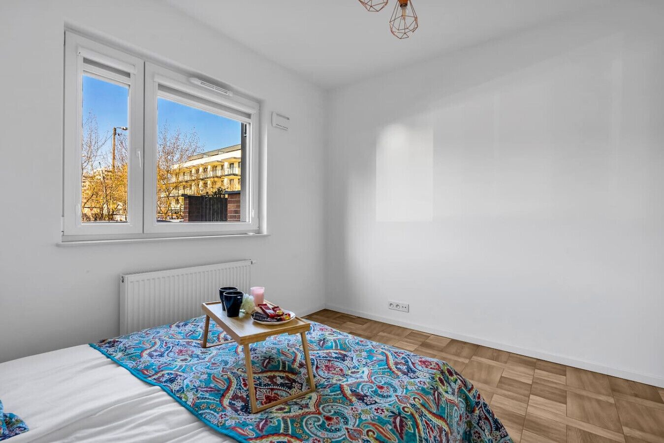 Ein Schlafzimmer mit Bett, einem Fenster mit Blick auf Gebäude, einem Heizkörper und einem kleinen Tisch mit Gegenständen darauf.
