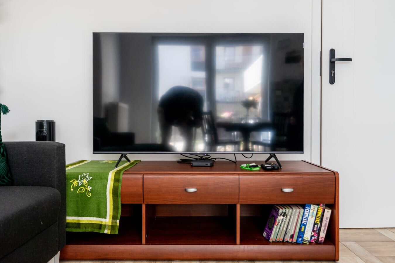 A flat-screen TV on a wooden TV stand with a green throw blanket, a remote, and a game controller. Books are stored in the lower shelf.
