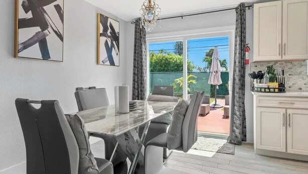 A modern dining area with a glass table and chairs, a chandelier, and artwork on the walls. - 4-bedroom house in charming Riviera Beach with WiFi, AC. Relax and unwind (RIVIERA BEACH)