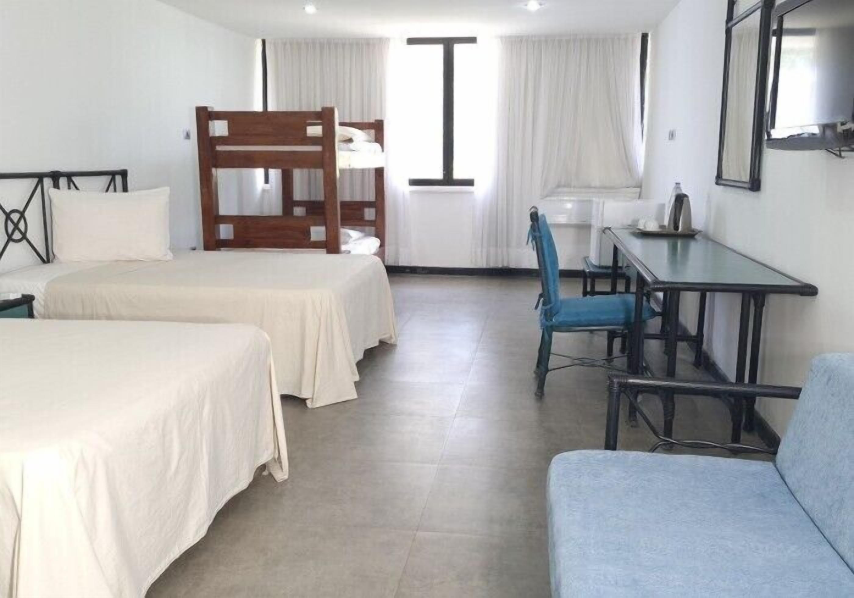 Quarto de hotel com duas camas, uma escrivaninha, uma cadeira e uma cama beliche.