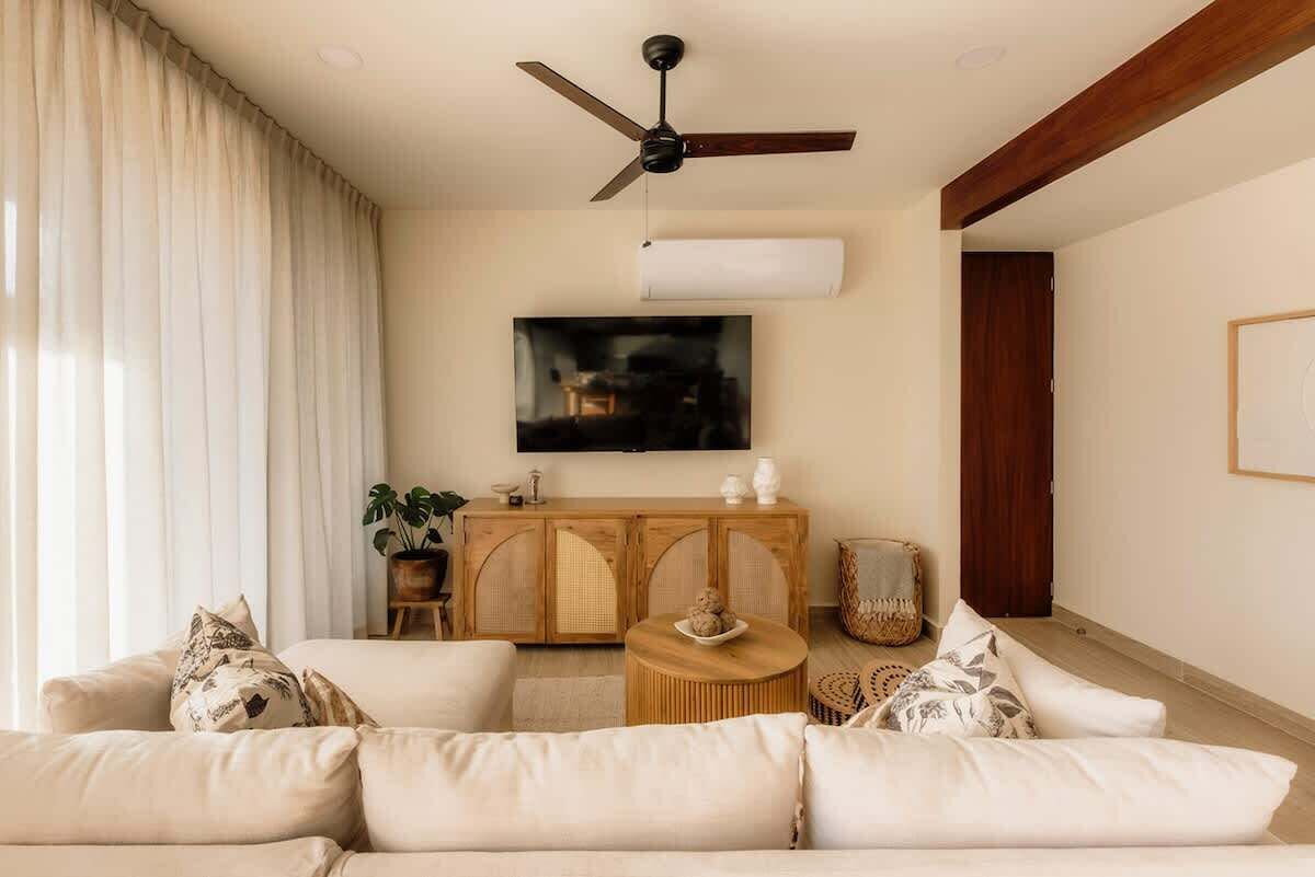 Uma sala de estar com sofá, ventilador de teto, televisão, aparador de madeira e uma planta.