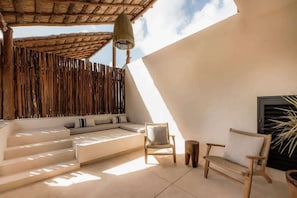 A sunlit outdoor seating area with a woven wall, a cushioned bench, and a wooden chair.