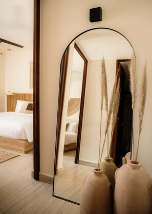 Un dormitorio con un espejo grande, una cama, una mesita de noche y dos jarrones con pastos altos.