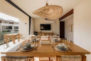 A dining area with a wooden table set for four, a woven ceiling light, and a view of a building outside.
