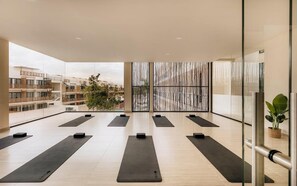 A spacious indoor area with multiple yoga mats arranged for use, large windows offering a view of the surrounding buildings, and a glass partition with vertical lines.