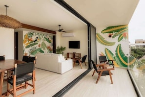 Uma sala de estar moderna com uma janela ampla, área de jantar com mobiliário de madeira e um mural com folhagens tropicais.