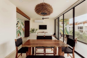 Uma sala de jantar com mesa e cadeiras de madeira, luminária de palha no teto, televisão de tela plana e vista para o exterior.