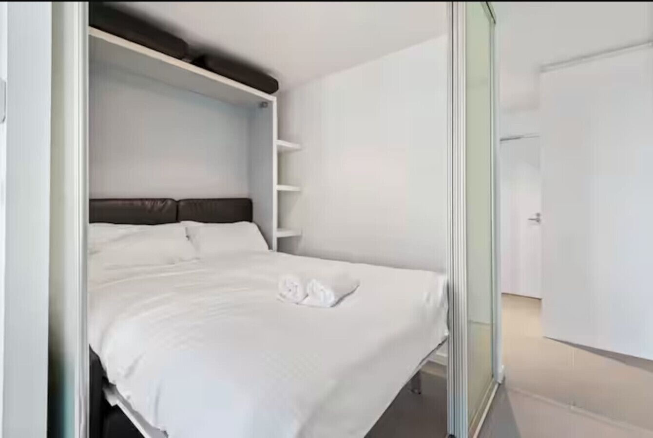 A neatly made bed with white linens and a folded towel, a dark headboard, and a glass door leading to a hallway.