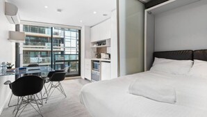 Une chambre d’hôtel moderne dotée d’un grand lit, d’un bureau avec des chaises, d’une kitchenette et d’un balcon offrant une vue sur la ville.