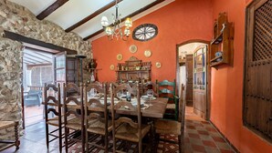 Un coin repas avec une table en bois dressée pour six personnes, entourée de chaises en osier. Un mur de pierre et des poutres en bois sont visibles sur la gauche, et une grande porte en bois ornée d’un motif en treillis se trouve sur la droite.