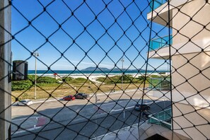 A view through a chain-link fence of a coastal road with vehicles, a beach, and a building with a blue facade. - Cf03 Apto vista mar da praia do forte (Cabo Frio)