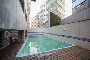 A swimming pool with a wooden deck, surrounded by high-rise buildings and a tiled wall with a decorative border. - Cf03 Apto vista mar da praia do forte (Cabo Frio)
