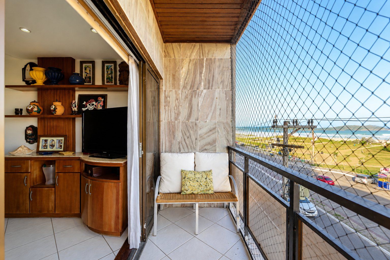 Un balcón con vista a la playa, una zona de estar con cojines y un televisor sobre un mueble de madera.