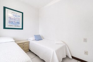 A bedroom with two beds, a nightstand, a framed picture of a cityscape, and a white wall. - Cf03 Apto vista mar da praia do forte (Cabo Frio)