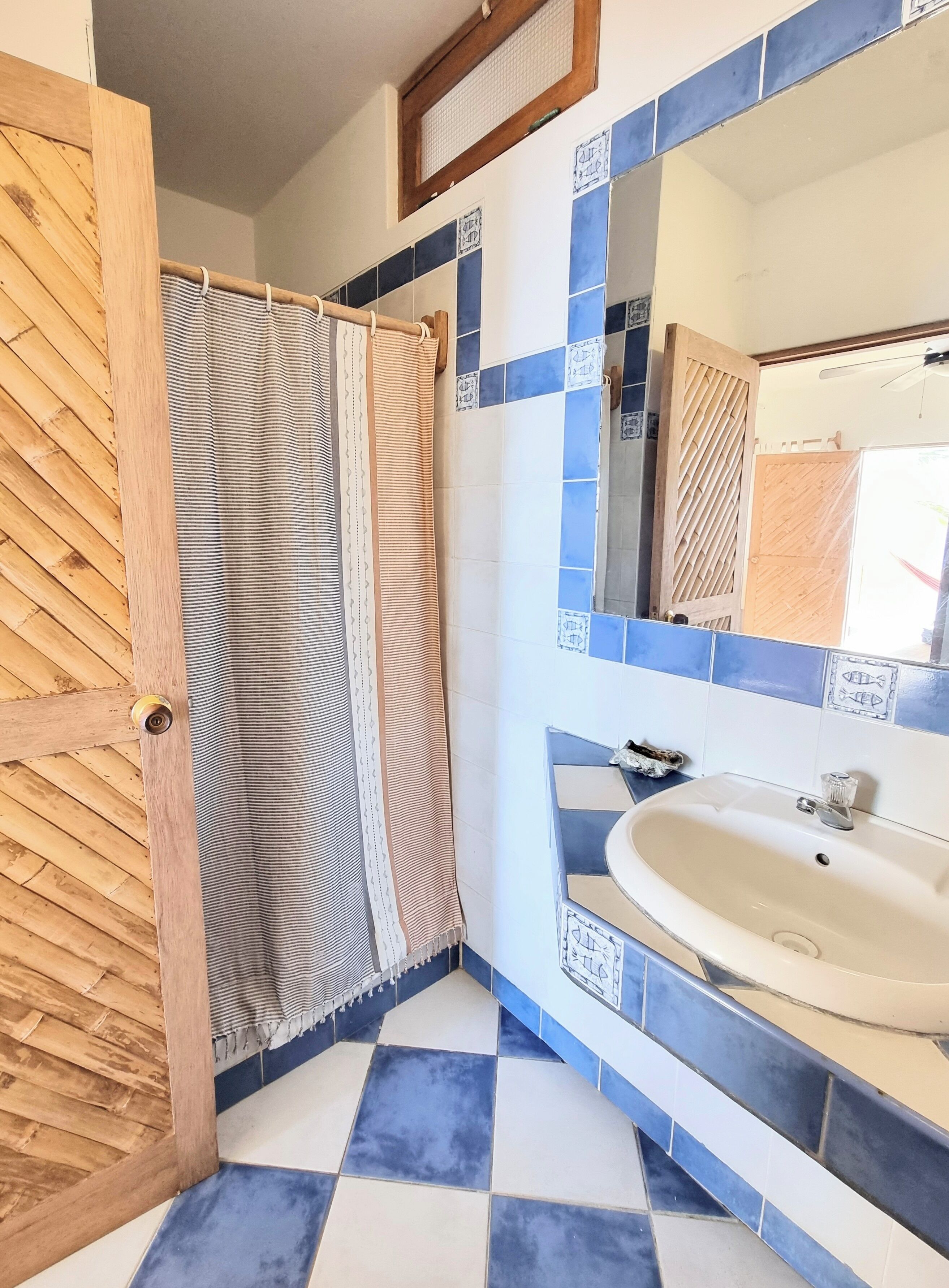 Un baño con suelo a cuadros, puerta de madera, cortina de ducha a rayas y un lavabo con espejo.