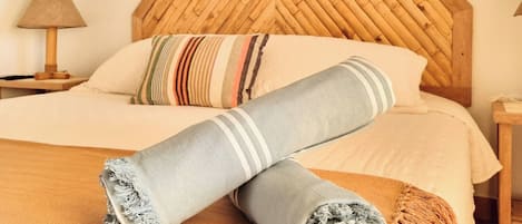 Una cama bien hecha con un almohadón a rayas, una manta enrollada y un cuadro colorido encima.