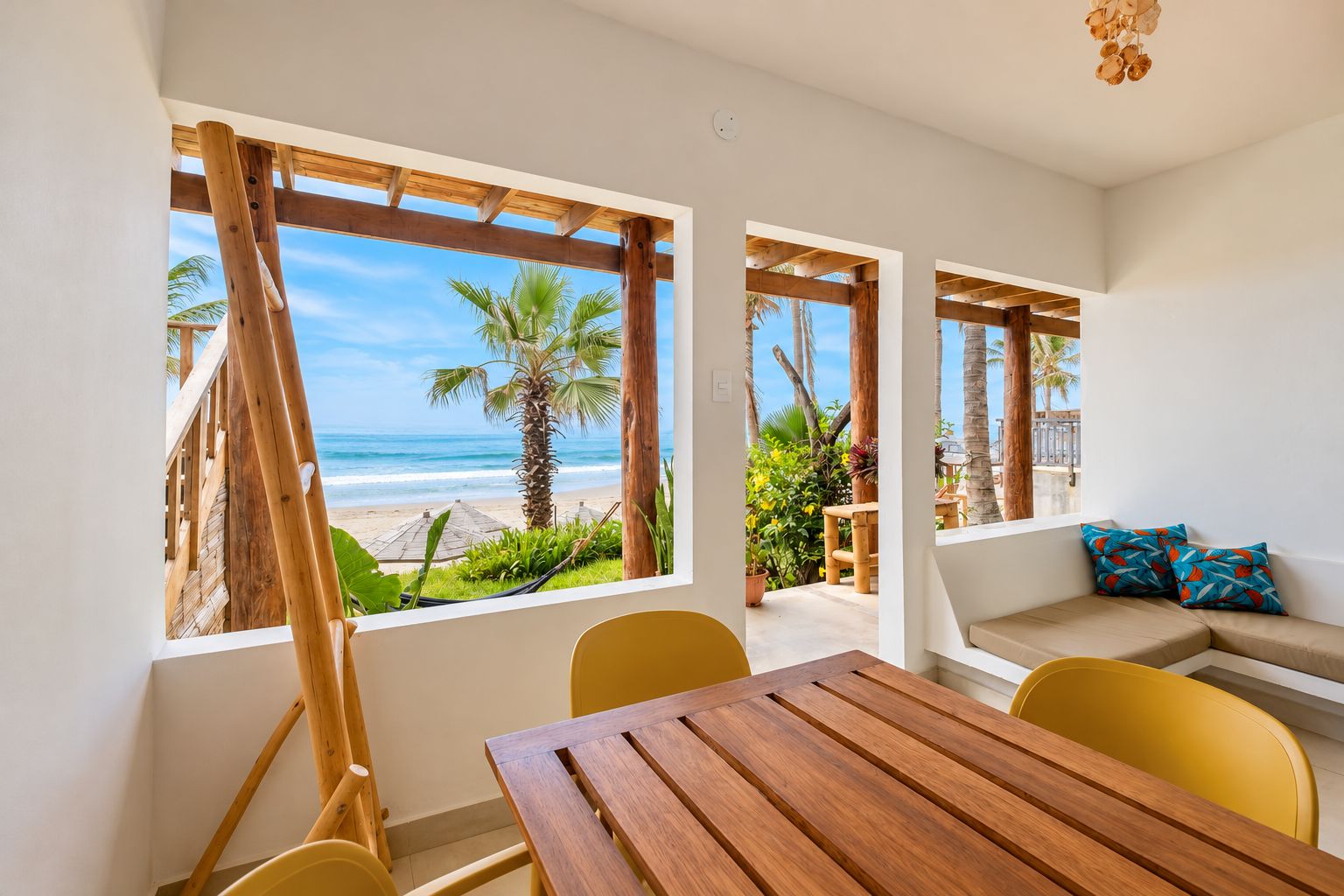 Ein Zimmer mit einem Holztisch und gelben Stühlen, ein beiges Sofa mit blauen Kissen und großen Fenstern mit Blick auf einen Strand mit Palmen.