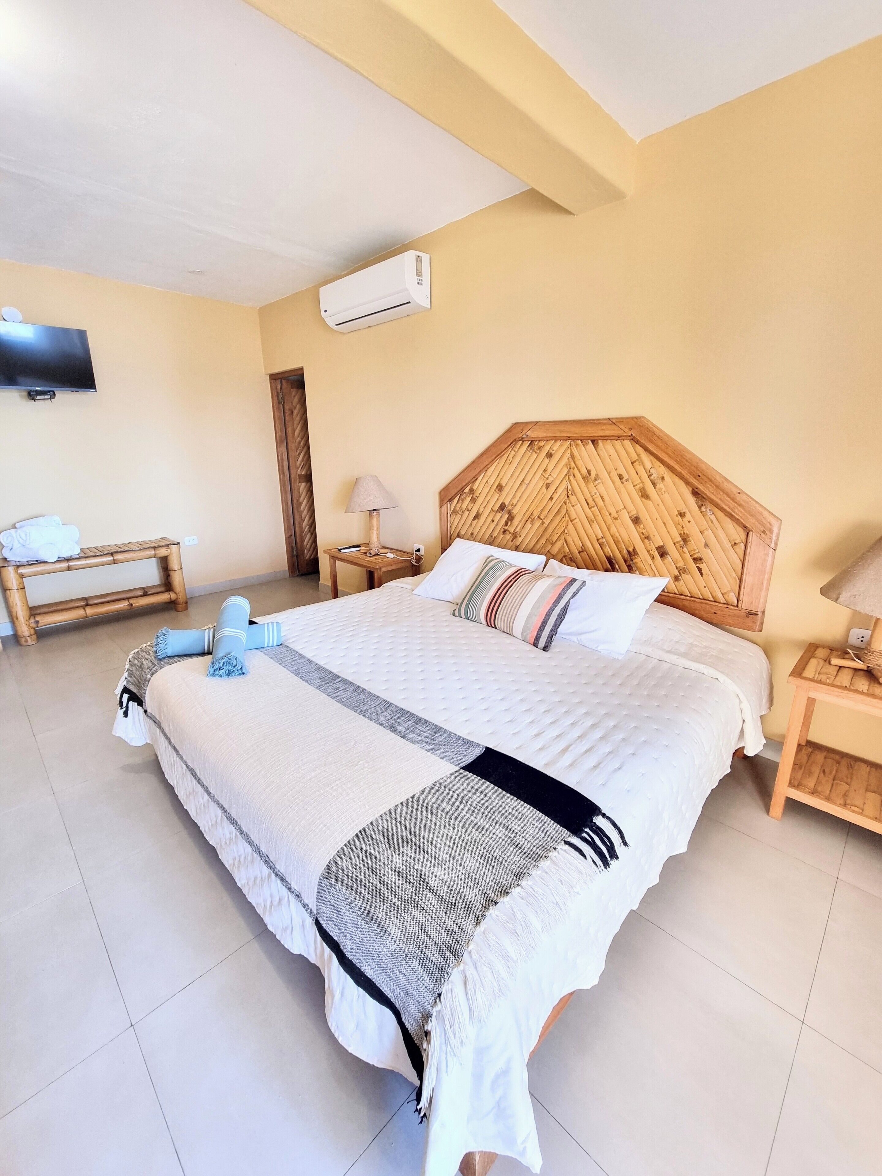 Una habitación de hotel con una cama grande, un televisor montado en la pared, un cabecero de madera y una mesita de noche con una lámpara.