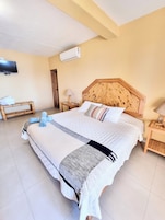 Una habitación de hotel con una cama grande, un televisor montado en la pared, un cabecero de madera y una mesita de noche con una lámpara.