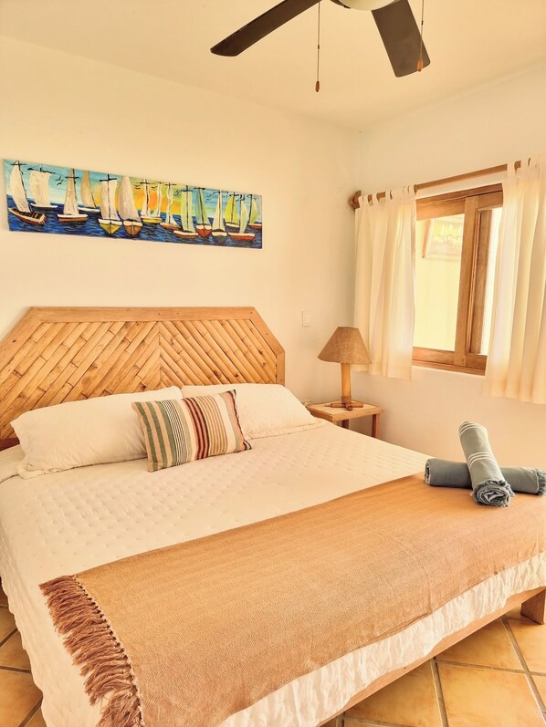 A bedroom with a wooden headboard, a painting of sailboats, a window with curtains, and a bed with a striped pillow and a rolled towel. - Mancora Beach Hotel (Máncora)