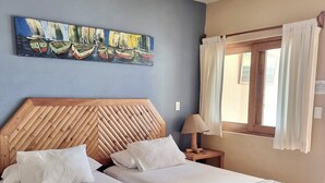 A double bed with a wooden headboard, two bedside tables, a ceiling fan, a window with curtains, and a painting of boats on the wall. - Mancora Beach Hotel  (Máncora)
