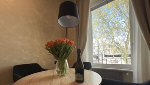 A round wooden table with a clear glass vase containing orange tulips and a bottle of Câdêl Bosco wine. The table is surrounded by black chairs, and there is a black lamp above it. A window with curtains is visible, showing a tree and a street view. - Pantheon Urbes Collection (Roma)
