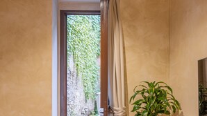 A hallway with a wooden floor, a potted plant, a bed, and a view of a garden through a door with curtains. - Pantheon Urbes Collection (Roma)
