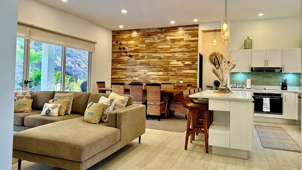 A modern living room with a sofa, dining area, kitchen, and a wooden accent wall. - 3-bedroom house Casa Tortuga (Coco)