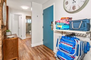 A hallway with a wooden floor, a blue door, a clock, and a shelf with folded blankets and a blue bag. - Tropical & Lush Paradise Vistas (Kihei)