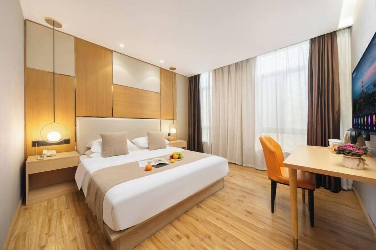 A modern hotel room with a large bed, wooden flooring, a desk with a chair, and a large window with curtains.