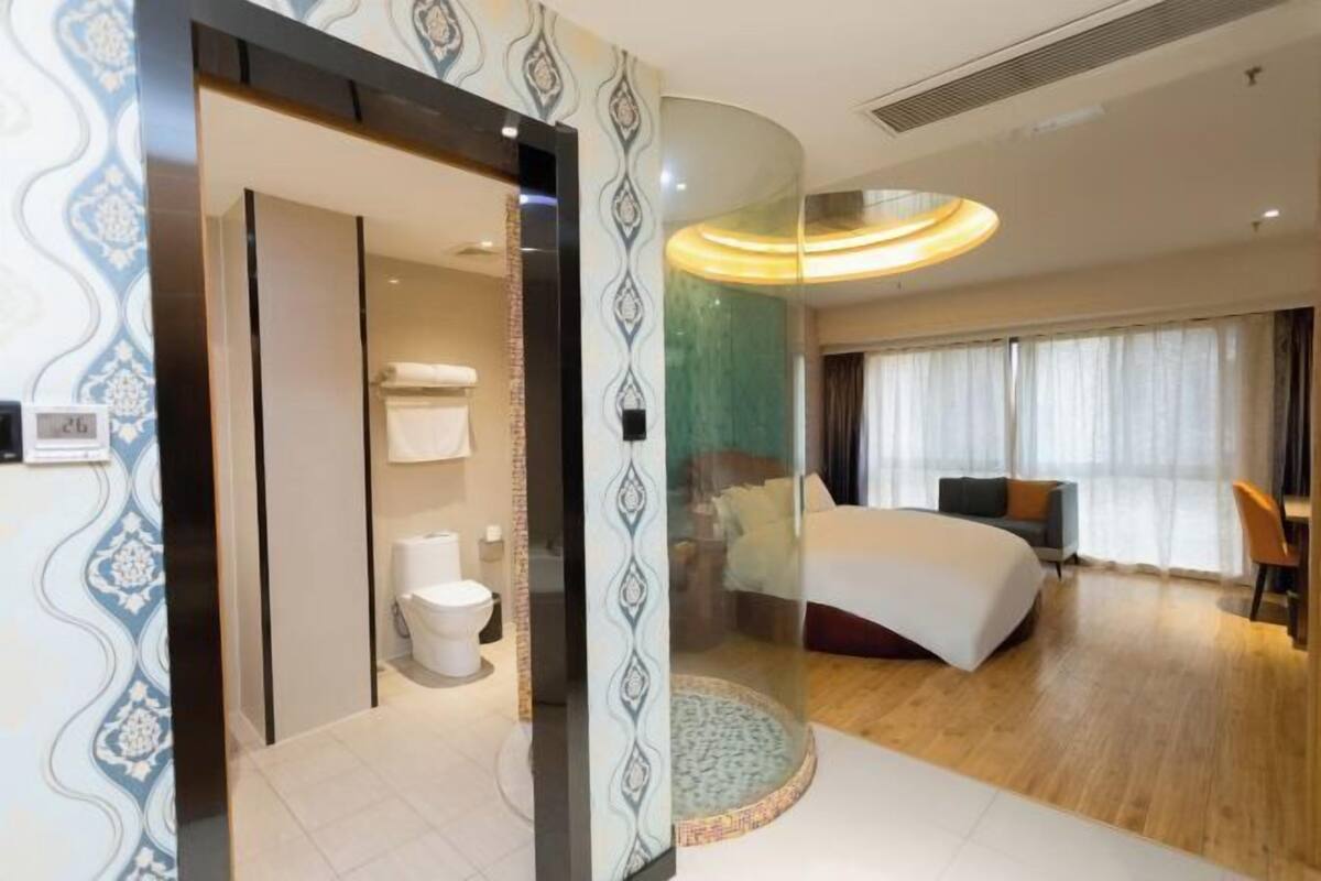 A hotel room with a bathroom, a bed, a round rug, and a view of the outside.