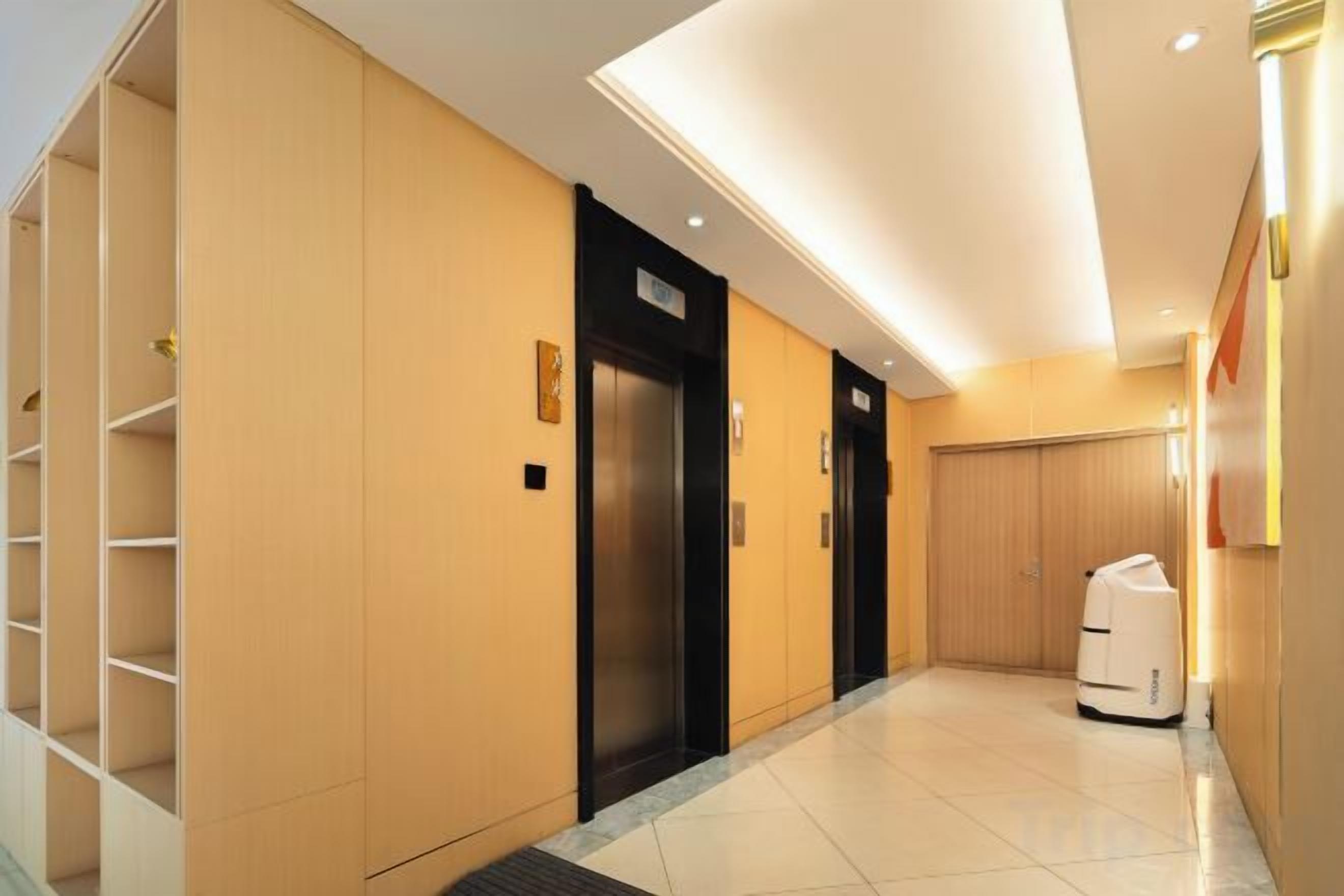 Een hotelgang met een modern design, met een centrale lift, een bagagekar en een houten deur.