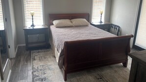Une chambre à coucher avec un lit en bois, deux tables de chevet avec des lampes, un tapis et des fenêtres équipées de stores.