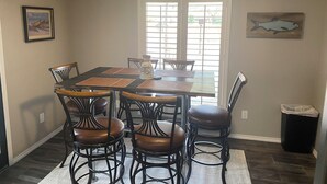 Un coin repas avec une table et des chaises, une fenêtre avec des stores et un tableau au mur.