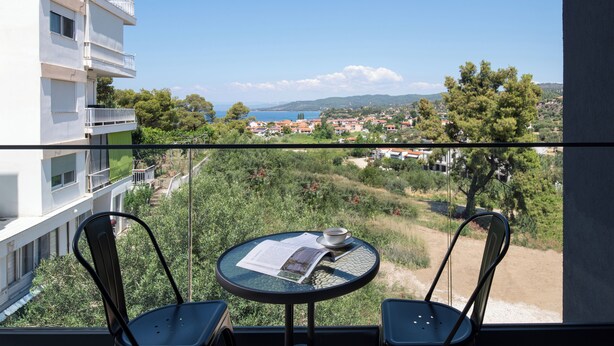 A balcony with two chairs, a table, and a view of a coastal town. - Acropol Golden Suites (Neos Marmaras)