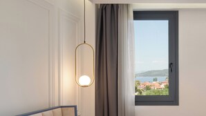 A hotel room with a bed, a bedside table, a hanging light fixture, a window with a view of a town, and a ceiling with recessed lighting. - Acropol Golden Suites (Neos Marmaras)