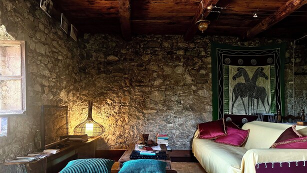 A rustic room with a stone wall, wooden furniture, a bed with a green tapestry, and a table with books and a lamp.