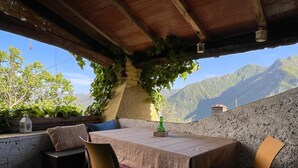 A rustic outdoor seating area with a table and chairs, surrounded by greenery and a mountainous backdrop.