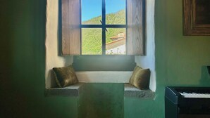 A room with a small window overlooking a green landscape, a piano, and a framed picture on the wall.