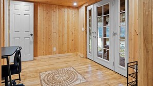 Una habitación con paredes de madera, una puerta blanca y un amplio ventanal que da al exterior. Hay una pequeña mesa y una silla en la esquina.