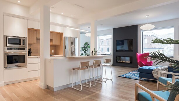 Un apartamento moderno con cocina, comedor y sala de estar. Incluye una cocina con electrodomésticos integrados, una mesa de comedor con taburetes tipo bar, una chimenea y un ventanal con vistas a la ciudad.