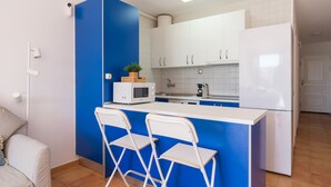 Cocina compacta con una pared de acento azul, armarios blancos, microondas, hervidor de agua y refrigerador. Hay dos taburetes blancos y una mesita pequeña.