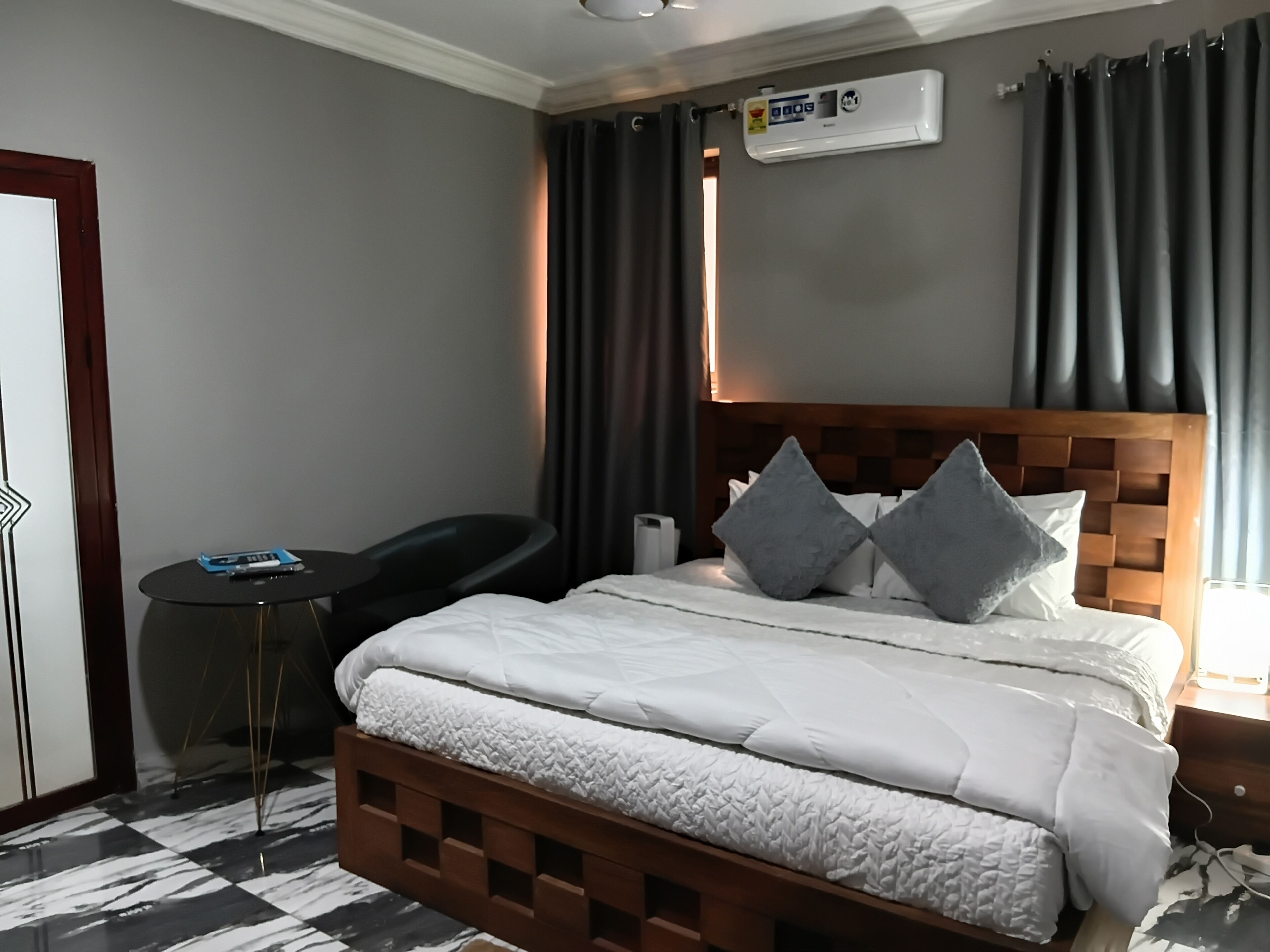 Habitación de hotel con cabecera de madera, cama con ropa blanca, dos almohadas, una mesita redonda pequeña y un aire acondicionado instalado en la pared.