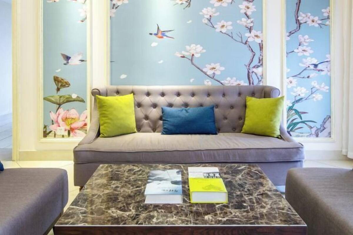 A living room with a grey sofa, two green and one blue pillow, a marble coffee table with books, and a wall with floral and bird wallpaper.