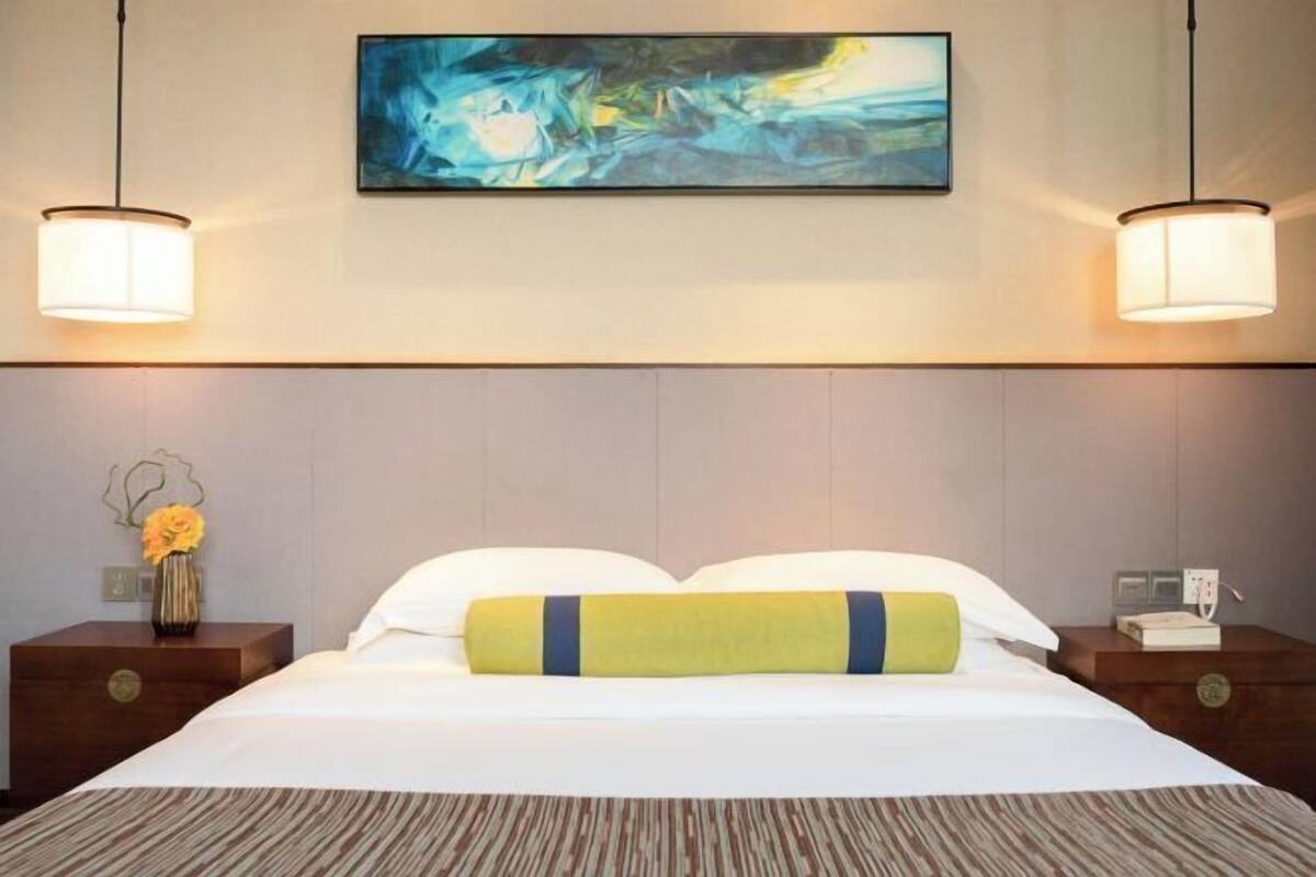 A hotel room with a large bed, two bedside tables, a nightstand, a vase with flowers, a painting on the wall, and two wall-mounted lamps.