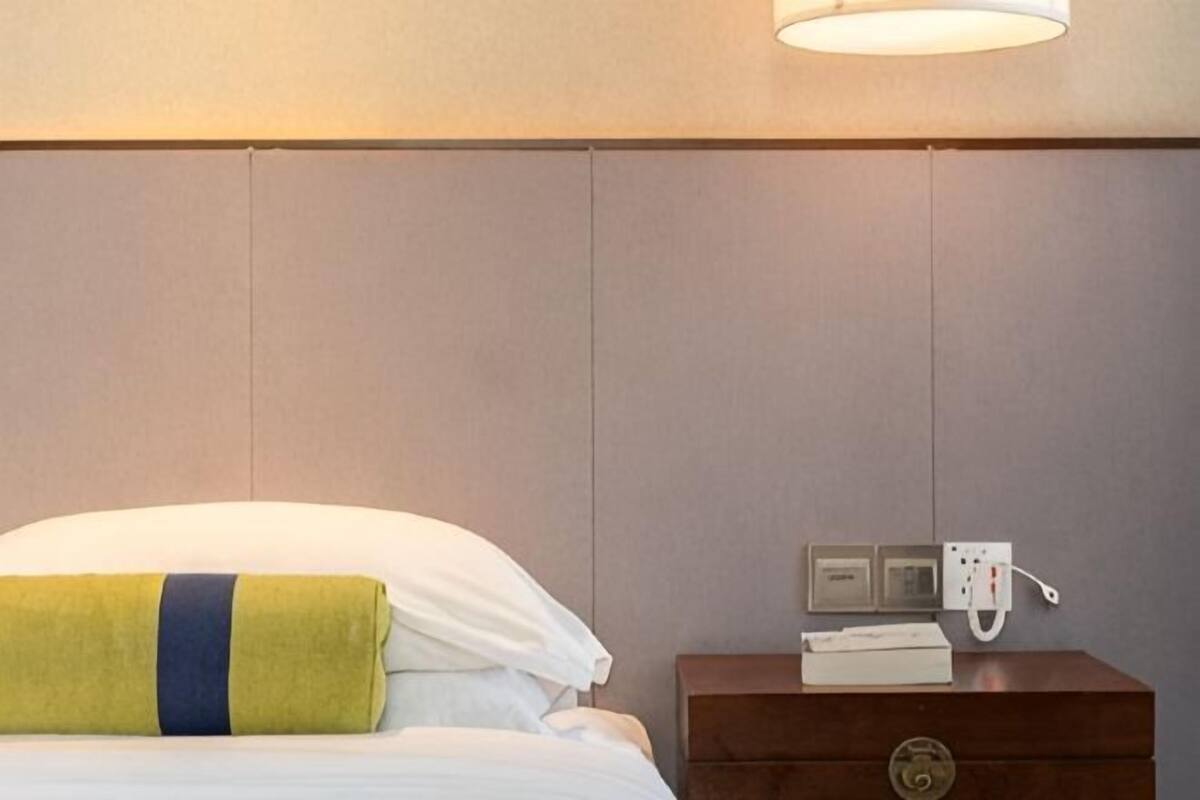A hotel room with a bed, a nightstand with a phone and a power outlet, and a wall-mounted light fixture.