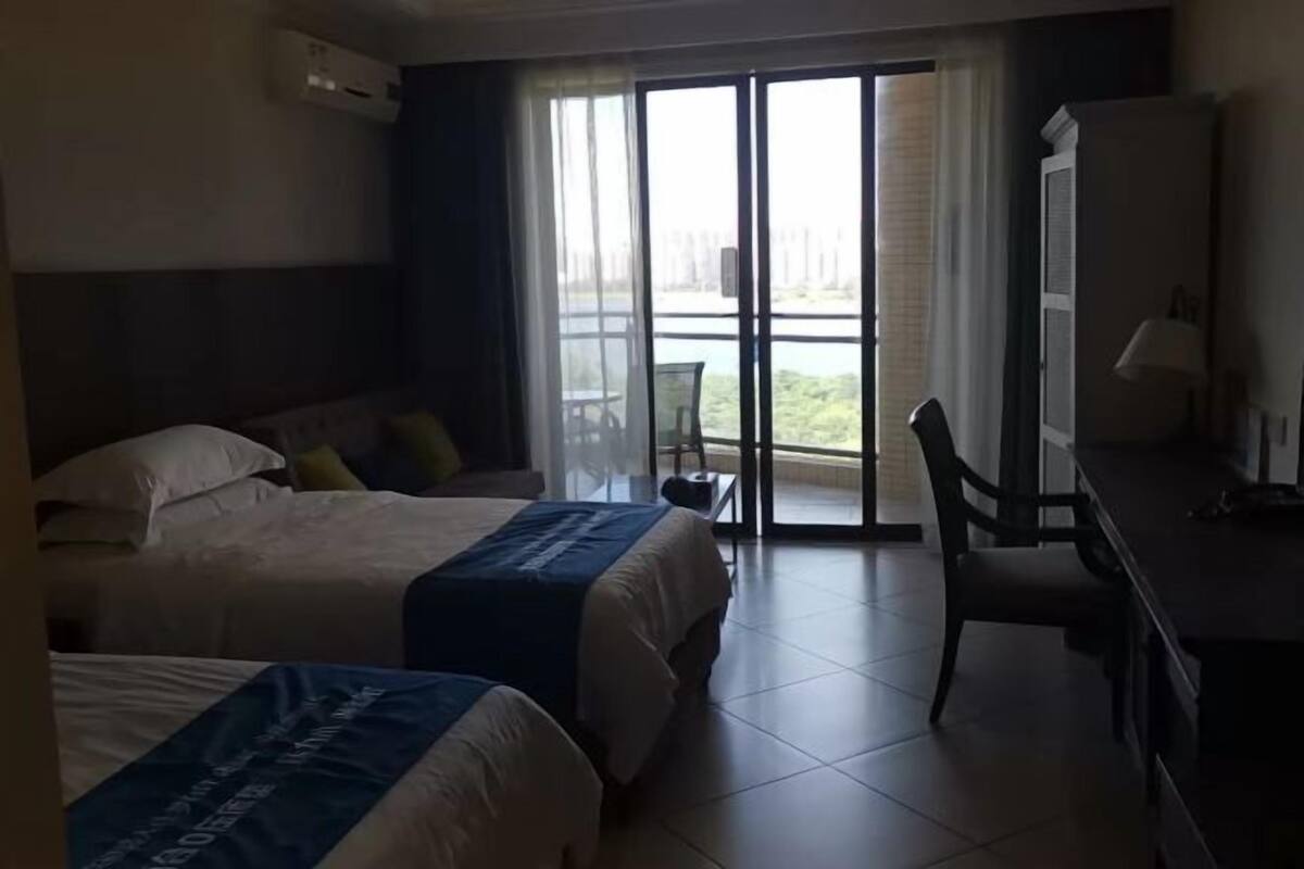 A hotel room with two beds, a desk, and a chair. There is a large window with a view of greenery and a building.