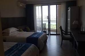 A hotel room with two beds, a desk, and a chair. There is a large window with a view of greenery and a building.