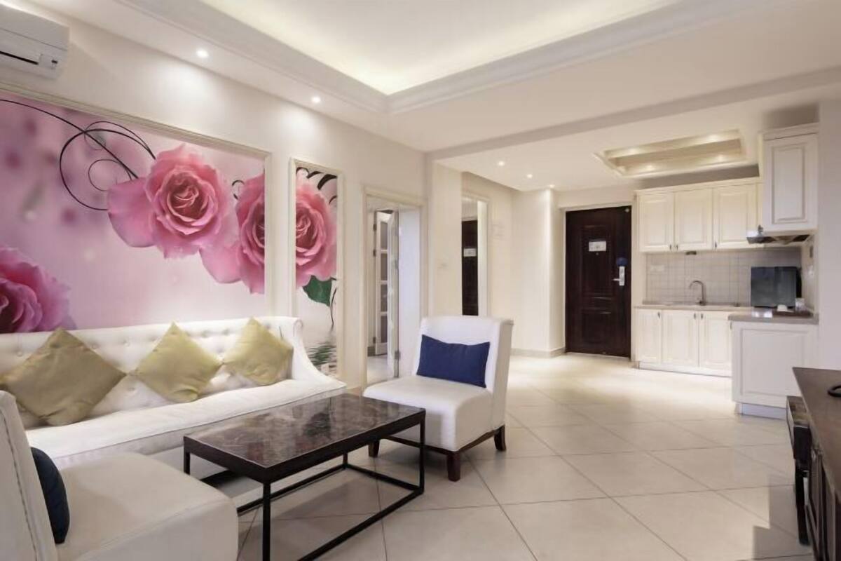A modern living room with a white sofa, a coffee table, and a chair with a blue cushion. There is a large floral mural on the wall. The room leads to a kitchen with white cabinets and a tiled backsplash.